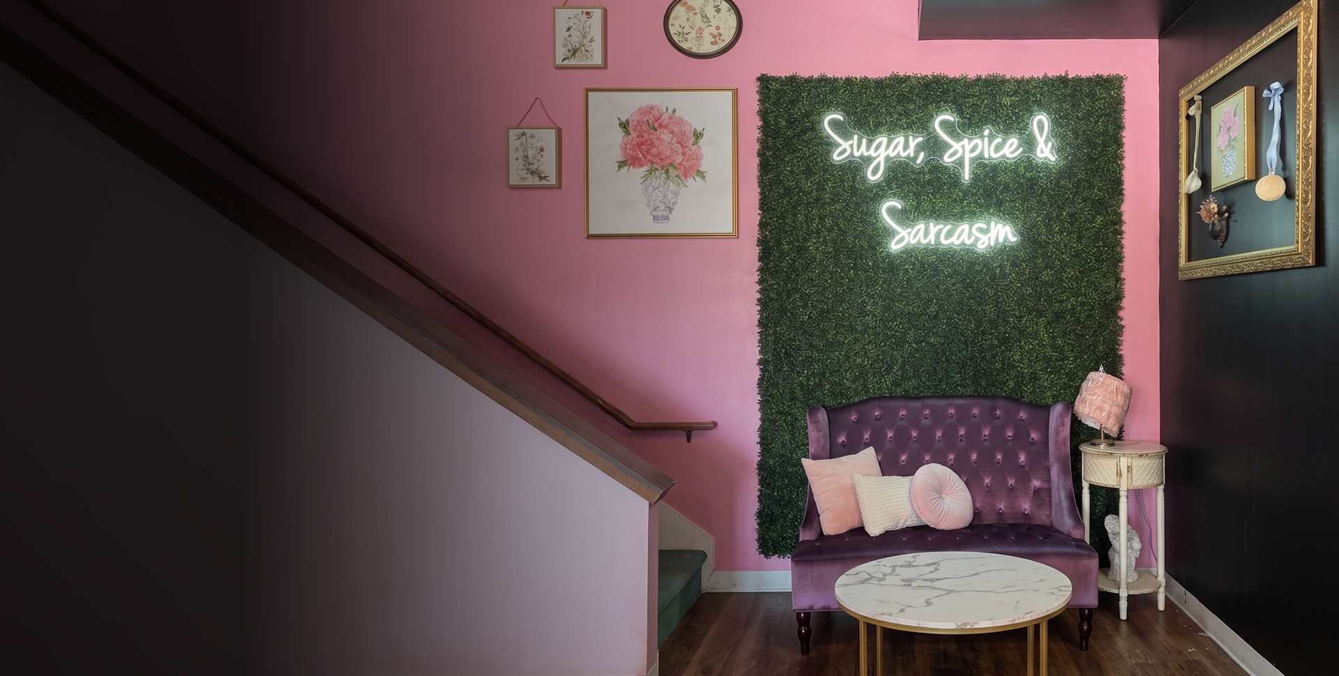 A cozy interior corner featuring a pink wall and a green hedge with a neon sign that reads 'Sugar, Spice & Sarcasm'. There is a purple velvet tufted loveseat with pink and white pillows, a small white side table with a pink lamp, and framed artwork on the wall, including floral prints and a textured piece with ribbons and small ornaments.