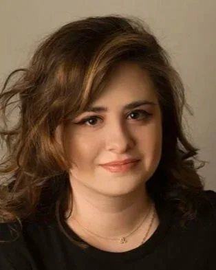 A woman with shoulder-length wavy brown hair, light makeup, and a small smile, wearing a black top and a delicate necklace, against a plain background.