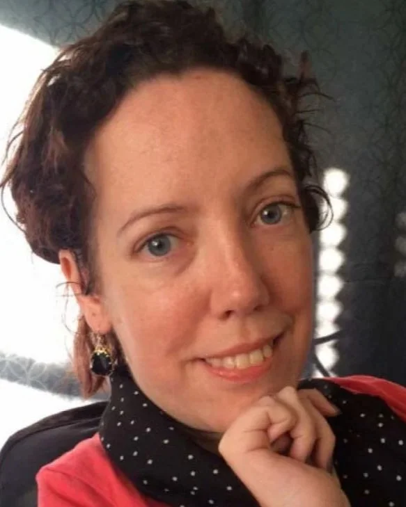 A woman with curly brown hair and blue eyes smiles, resting her chin on her hand, wearing a black and white polka-dot scarf and a red top, with soft lighting and a patterned wall in the background.