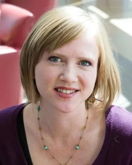 A woman with blonde hair and blue eyes smiling, wearing a purple top and a necklace, sitting near a window with sunlight.