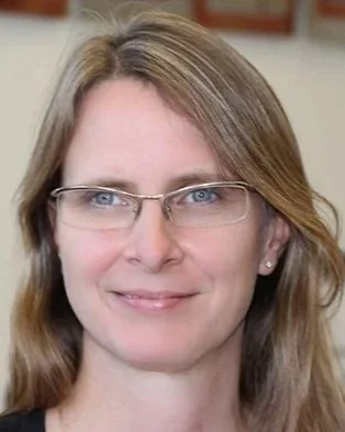 A woman with shoulder-length blonde hair wearing glasses, smiling, indoor setting.