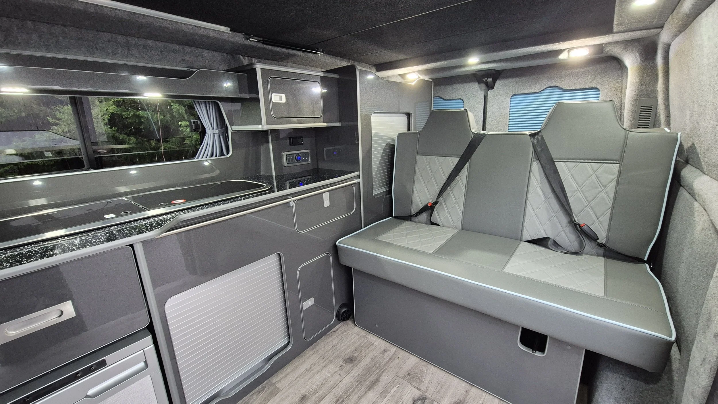 Interior of a modern camper van with a seating area and a compact kitchen, featuring a seat with black seat belts, small cabinets, and a window with curtains.