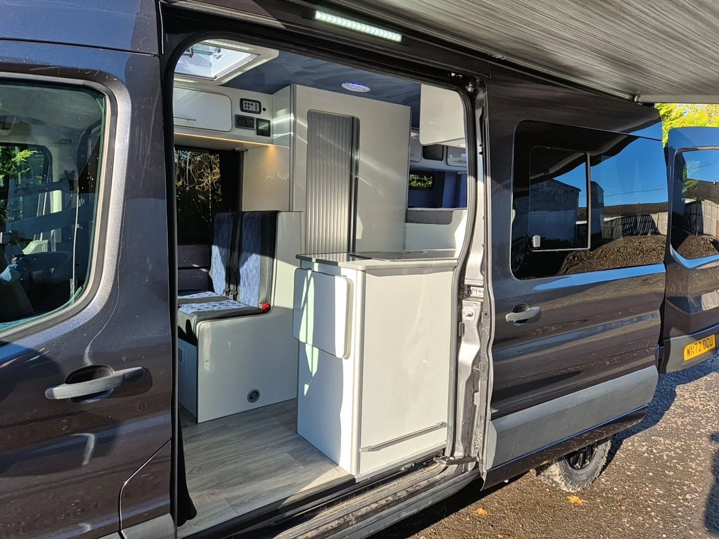 Inside a parked van showing custom cabinetry, a small refrigerator, and seating area with a window, with part of the van's exterior and a residential area visible.