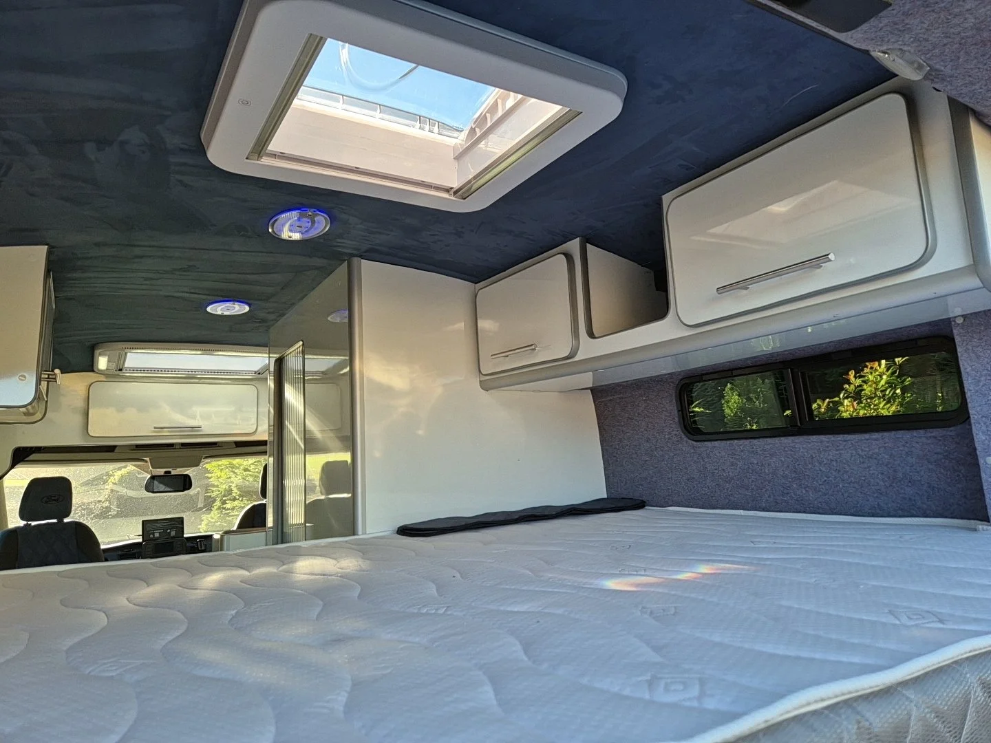 Interior of a camper van showing a bed with a mattress, overhead storage compartments, a small window, and the dashboard area with two front seats visible.