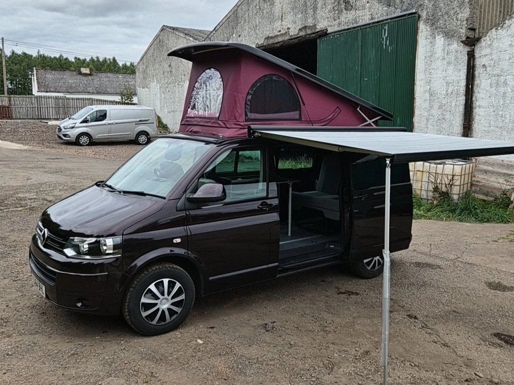 A black van with a pop-up tent on the roof and an extendable awning on the side, parked in a rural area near a barn.