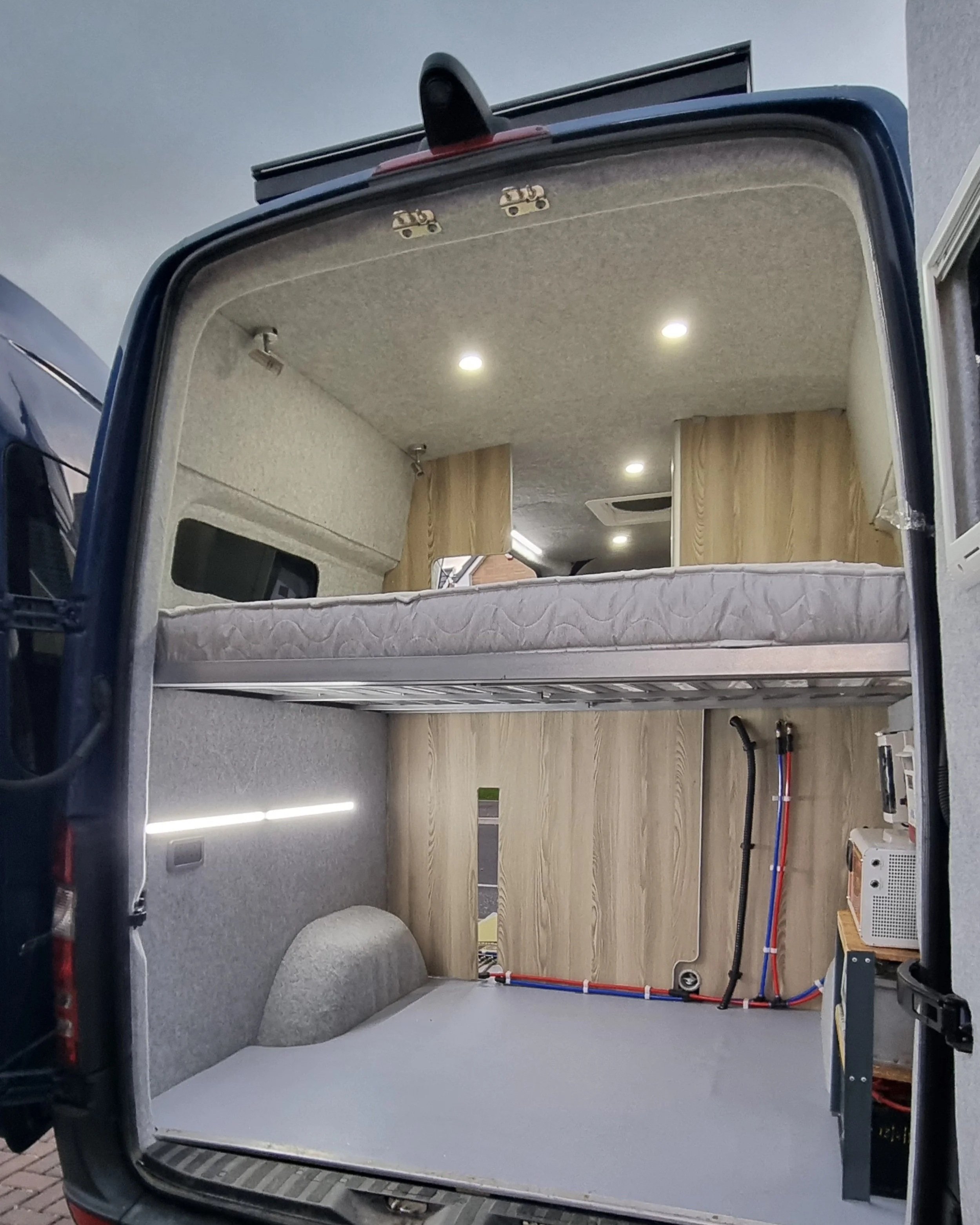 Interior of a camper van with a raised bed and wooden cabinets, LED lighting, and exposed electrical wiring.