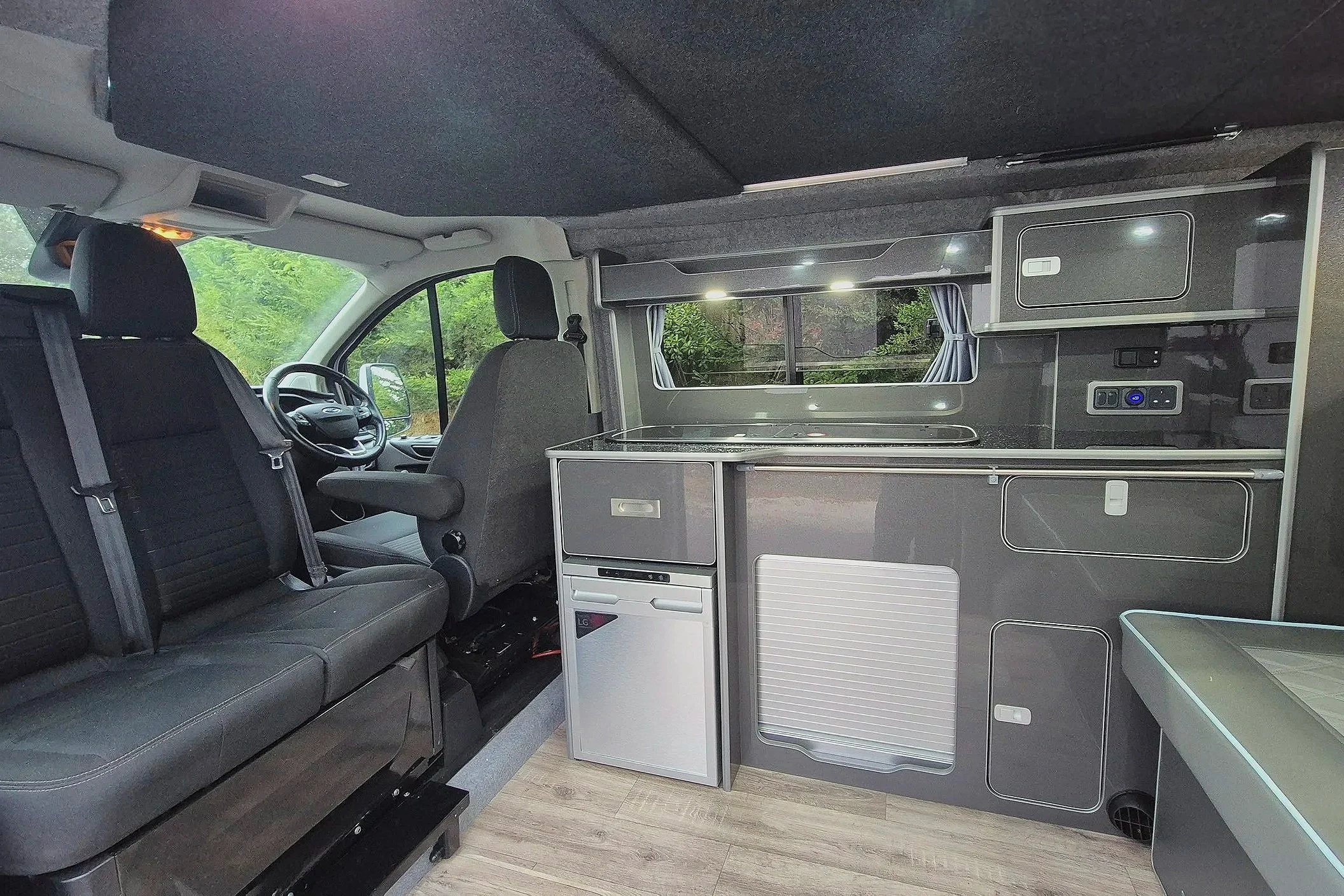 Interior of a camper van showing the front driving area with two seats and a compact kitchen area with cabinets, a small fridge, and a window