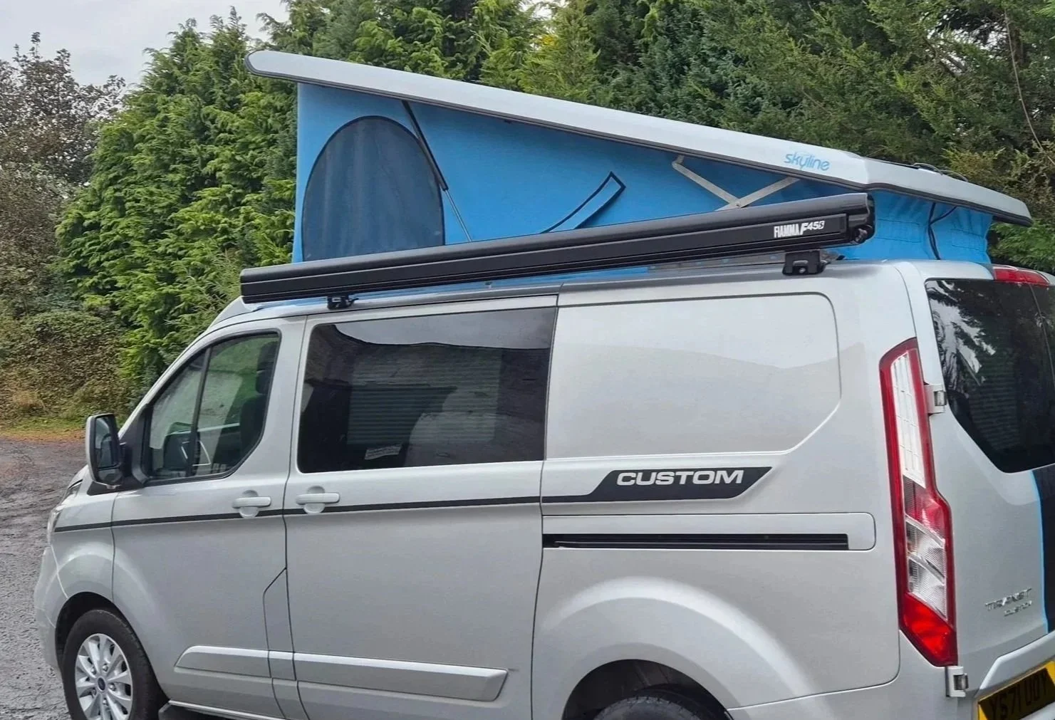 Silver camper van with a blue pop-up roof tent, a black rooftop cargo box labeled 'Rhino F445,' parked outdoors with greenery in the background.