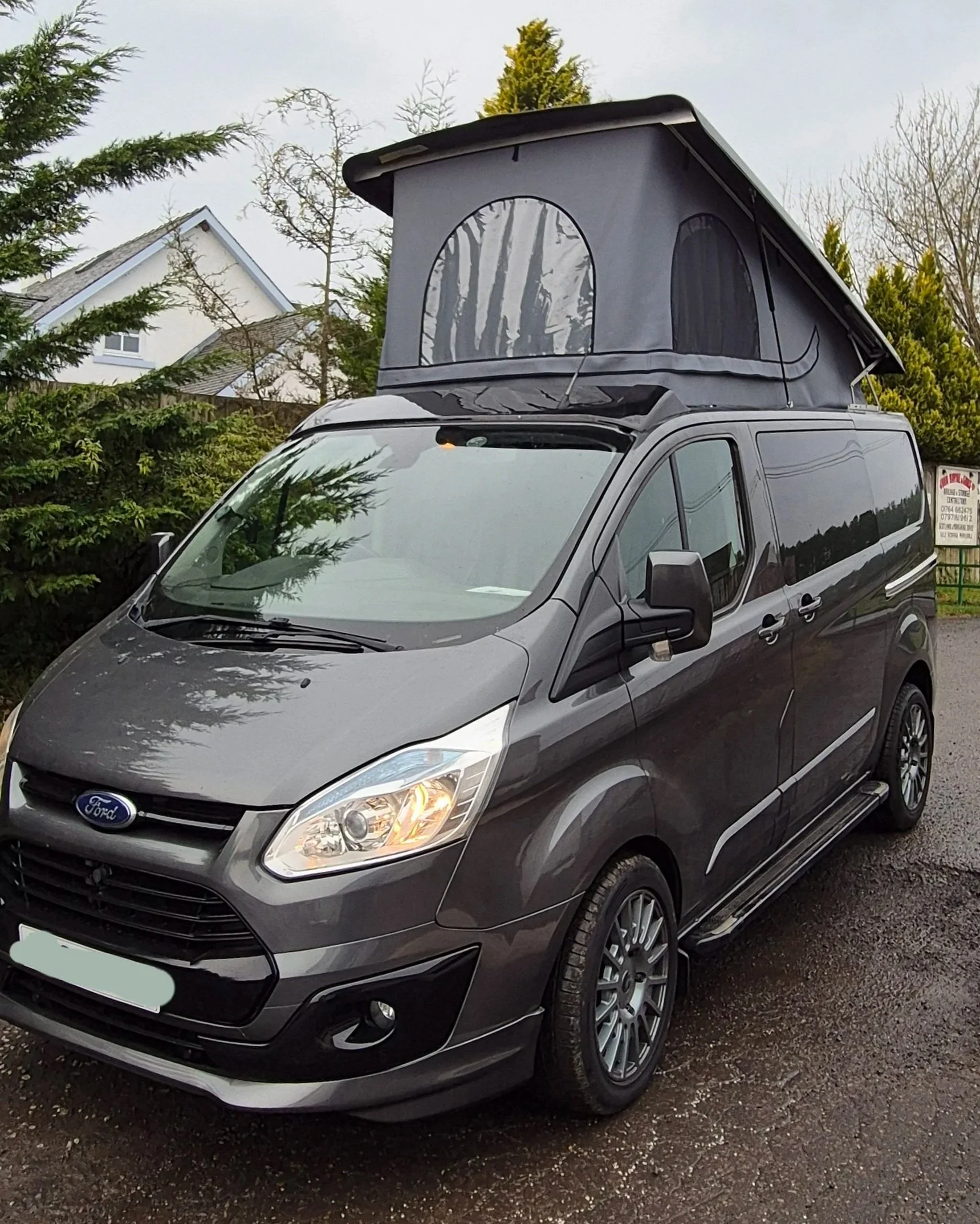 Gray Ford camper van with a raised rooftop tent parked on the street, with trees and houses in the background.
