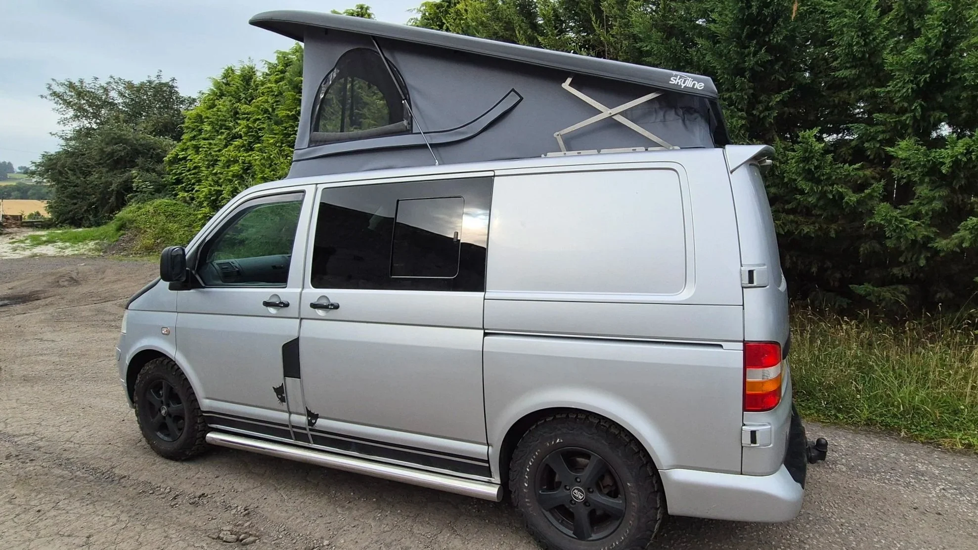 A silver camper van parked outdoors on a dirt surface, with a pop-up roof tent extended on top and surrounded by green trees and grassy areas.