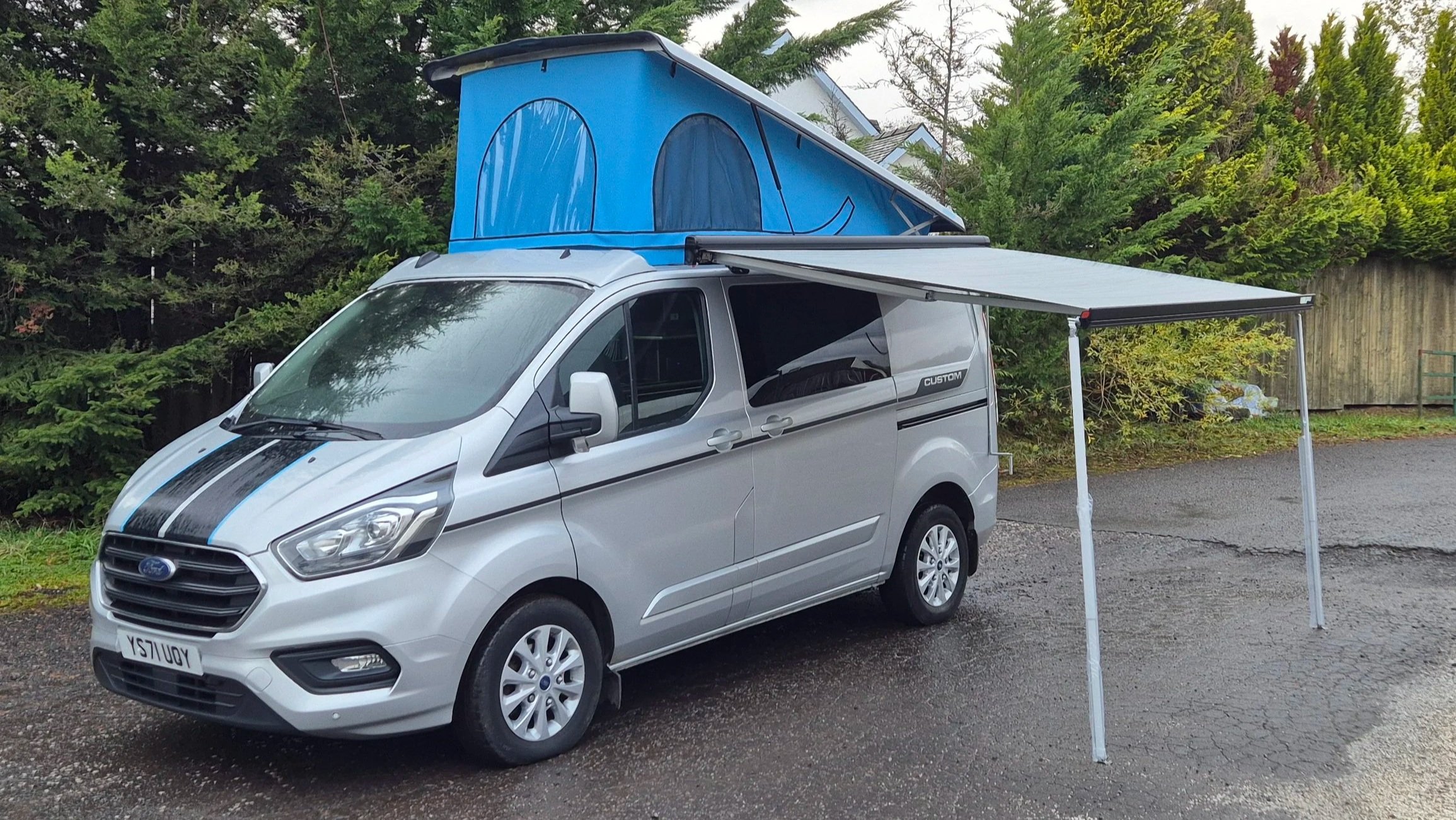 Silver Ford Transit van with custom black racing stripes, a blue rooftop tent, and an extended fabric awning on a wet, paved surface near green trees and a wooden fence.