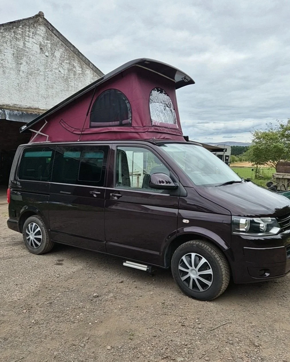 A black camper van with a pink elevated tent on top, parked outdoors near farm buildings and trees under cloudy skies.