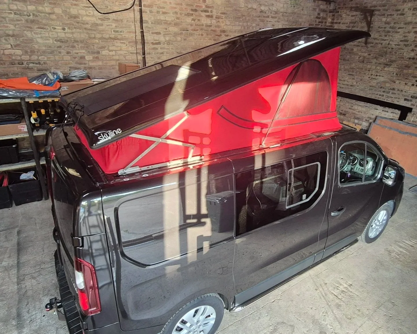 Black van with a red roof tent set up on top inside a warehouse with brick walls.