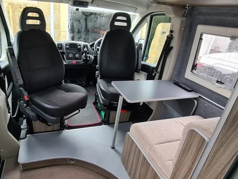 Inside a van converted into a camper or mobile home with two driver seats at the front, a small fold-out table, a cushioned seat, and a window on the side wall.