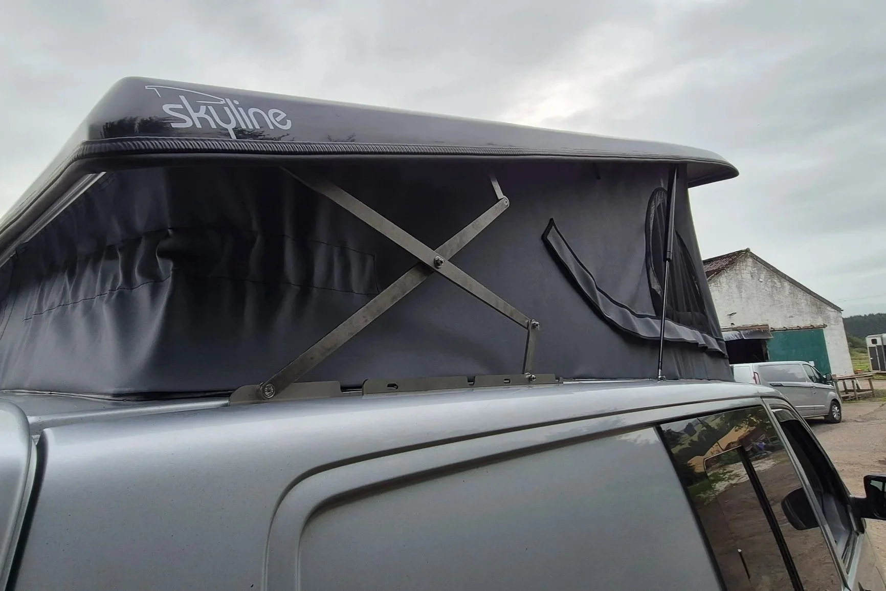 A silver van with a black Skyline rooftop tent mounted on top, partially extended with metal supports visible, set outdoors against a cloudy sky and a rustic building in the background.