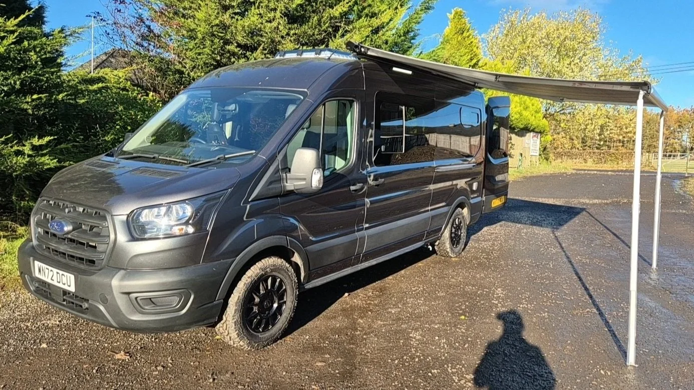 A black camper van with a retractable awning extended on the right side, parked on an unpaved area surrounded by green trees and blue sky.