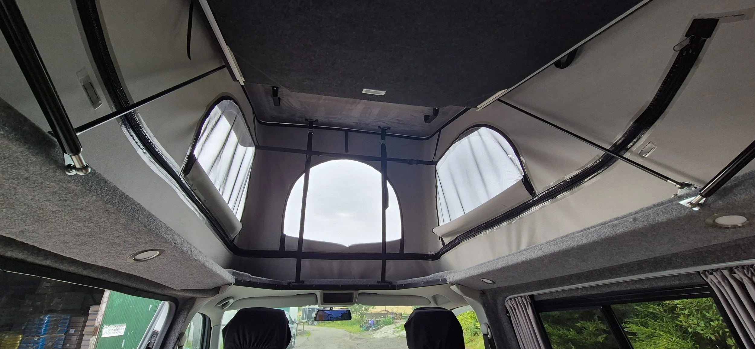 Inside view of a camper van showing the ceiling with a large open window, fabric curtains, and structural support bars.