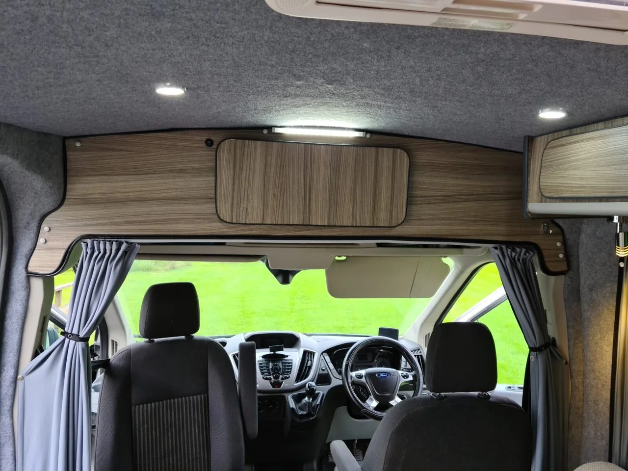 Interior of a camper van showing the front seats, dashboard, steering wheel, and overhead wooden cabinets with a small light.