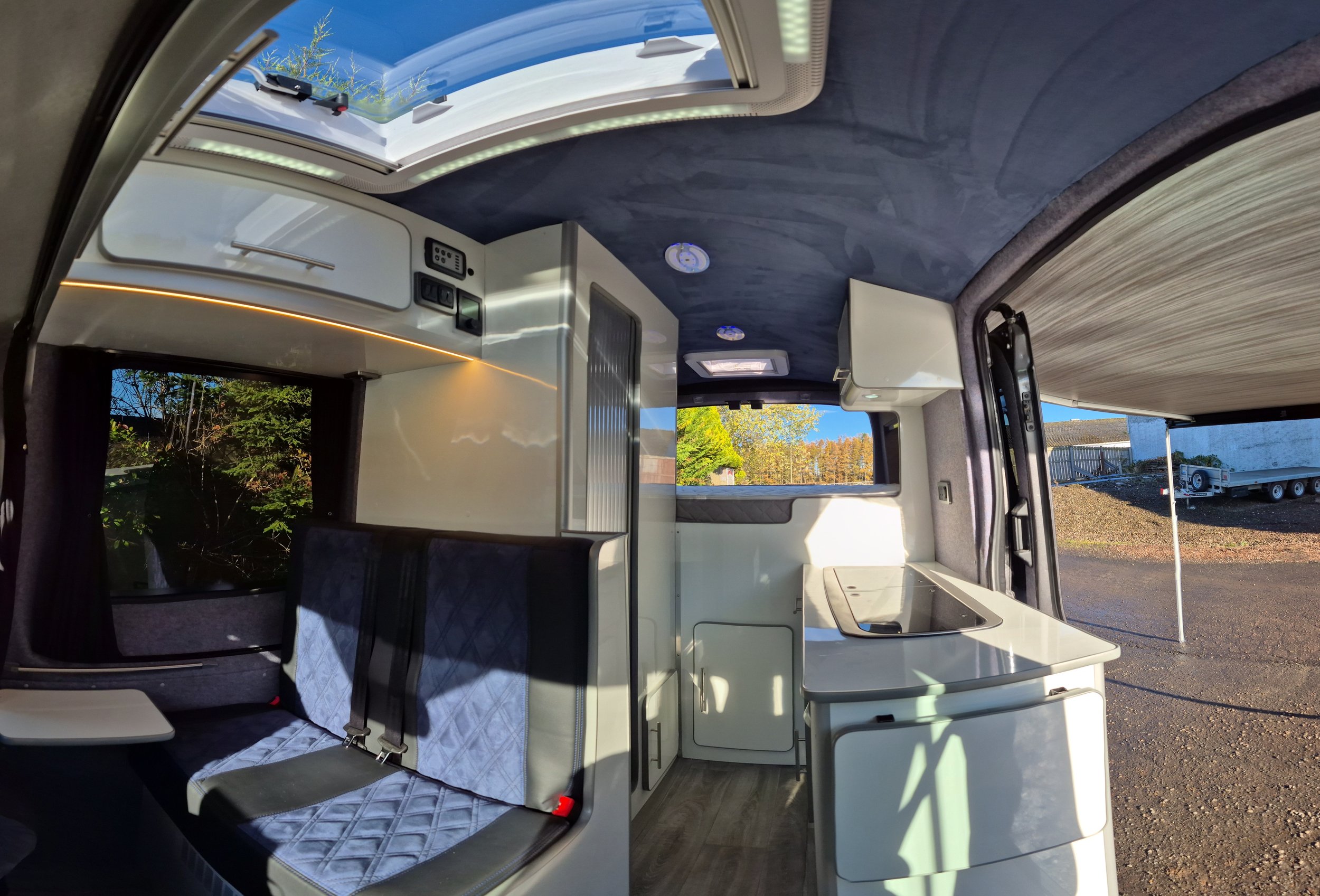 Interior of a camper van showing a sitting area with a bench seat, a small kitchen with a sink, and a skylight in the ceiling. Outside, there are trees and a trailer visible through the windows.