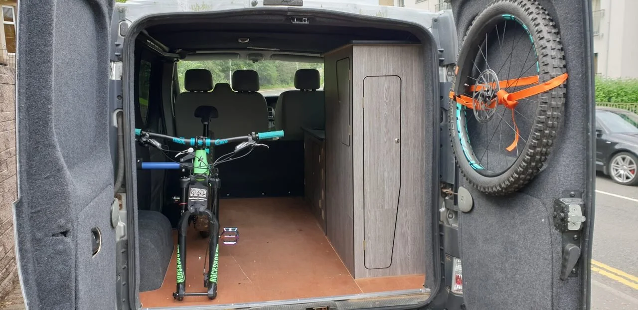 A van interior converted with a wooden cabinet and a mini bike, with a mounted bicycle on the door's side, parking lot visible outside.