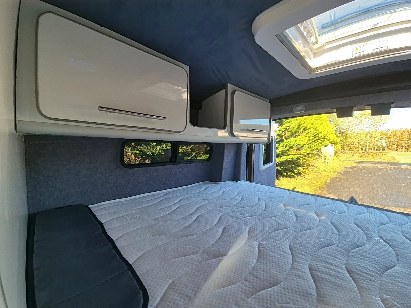Interior view of a camper van or RV with a bed, built-in storage cabinets, window, and a sky light, with trees and grass visible outside.