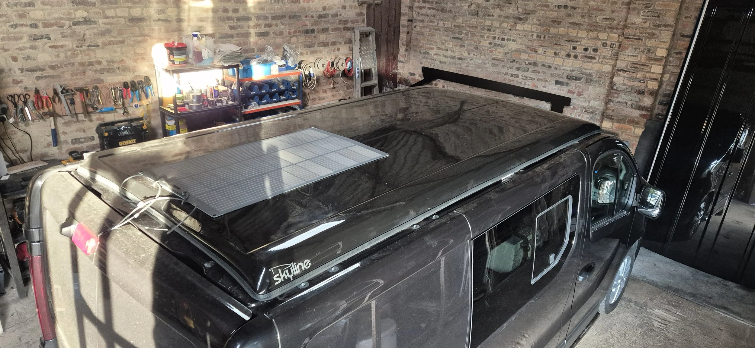 A black van with a solar panel on its roof parked inside a garage with brick walls, a workbench with tools and supplies, and various equipment along the wall.
