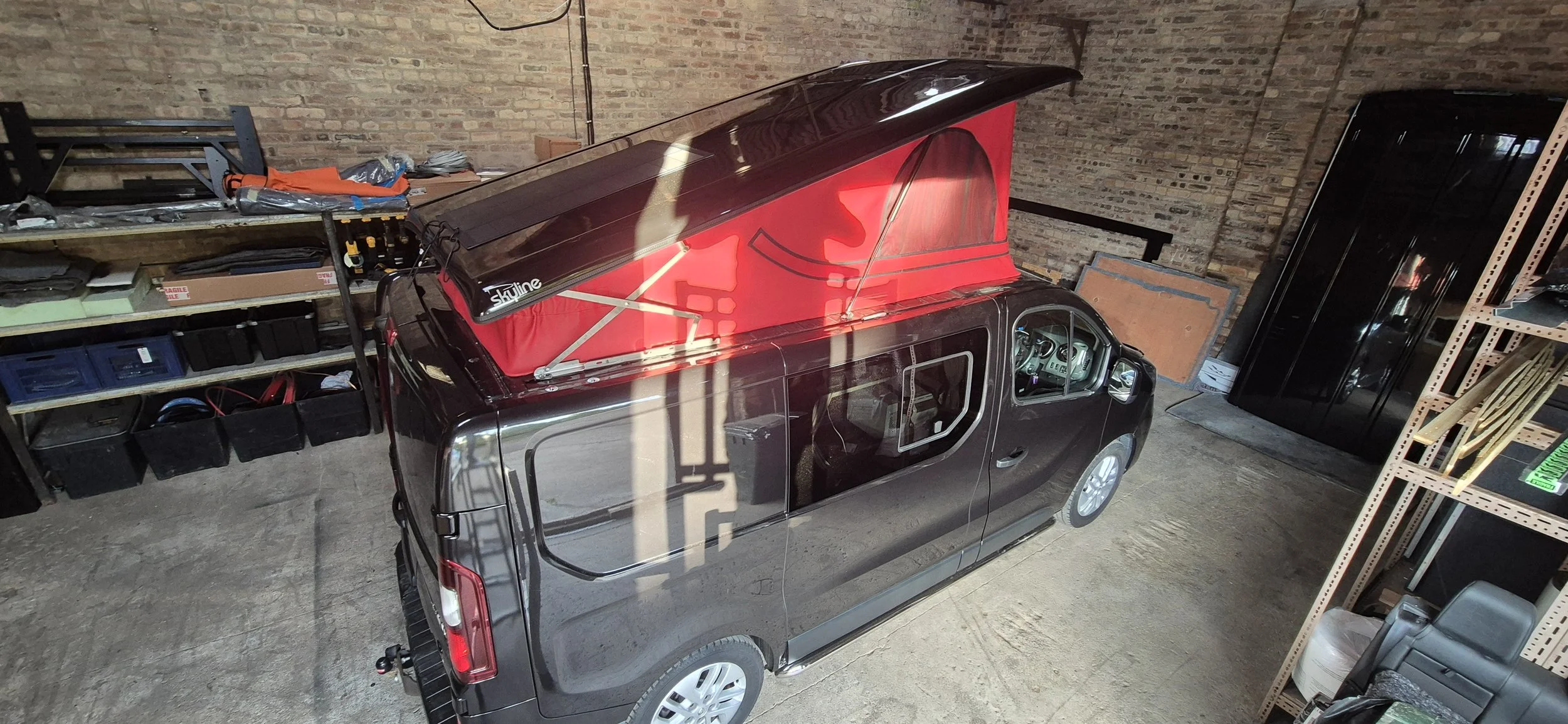 Black vehicle with a red rooftop tent set up inside a garage with brick walls and storage shelves.