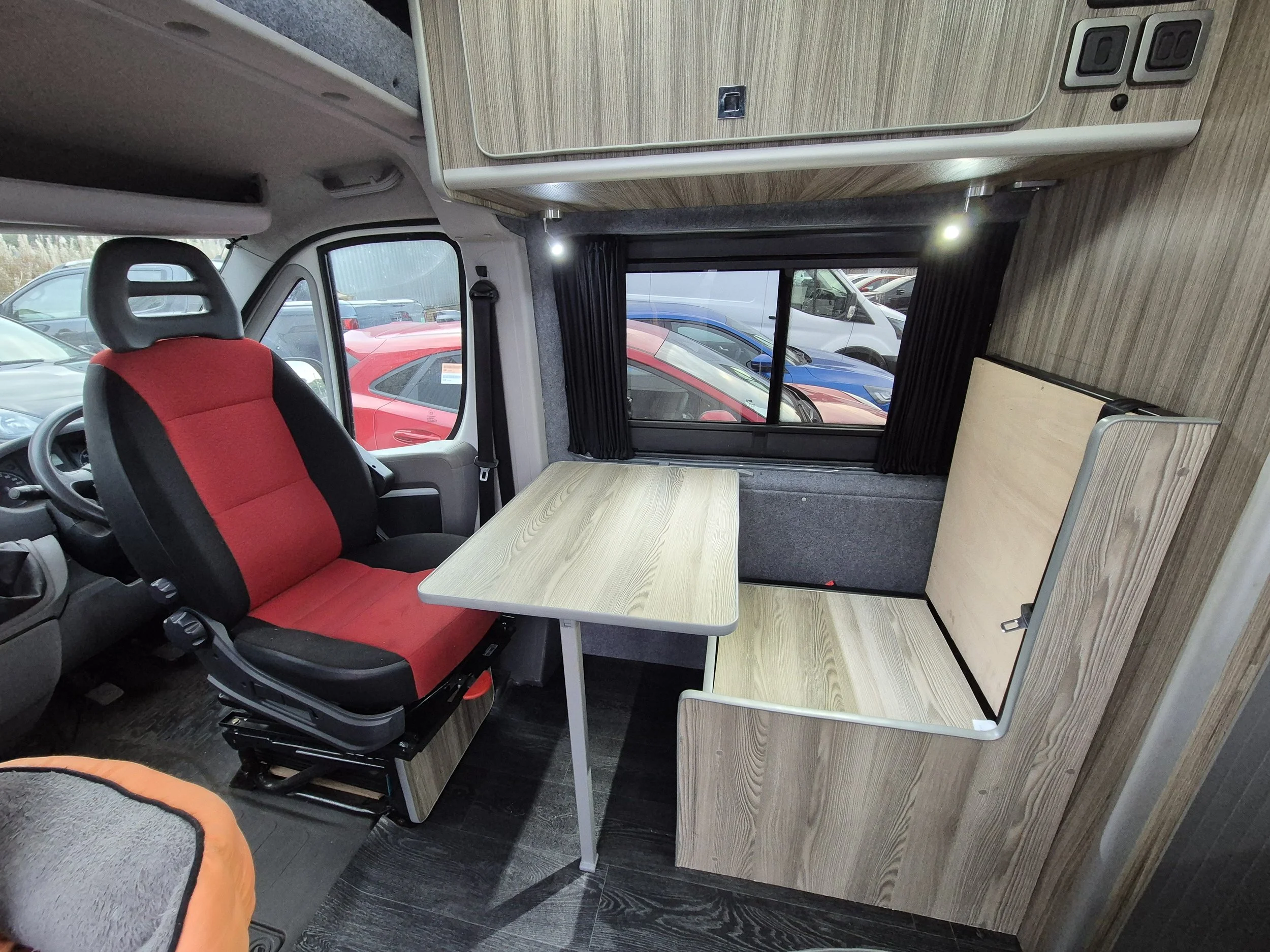 Interior of a camper van with a driver's seat, a small wooden table, a window with black curtains, and foldable seating or bed area in a compact, wood-paneled space.