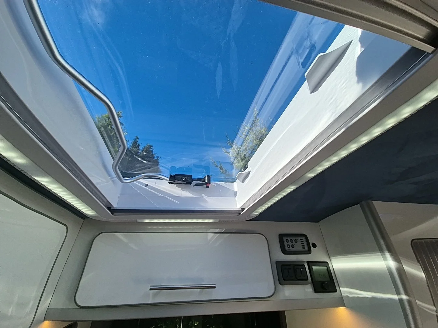 View through a skylight showing trees and a blue sky outside, with control panels and a vent inside the vehicle.