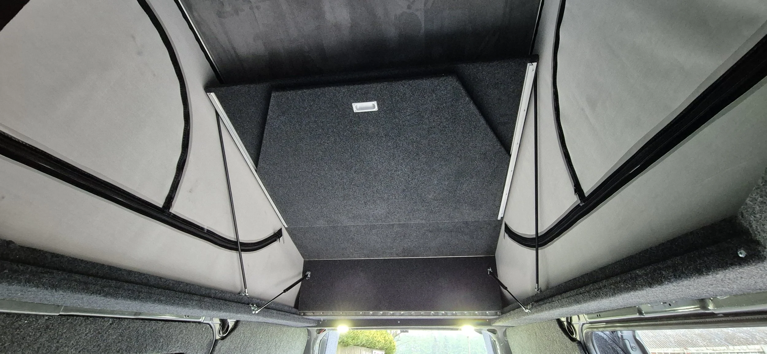 The interior of a pop-up camper showing the sleeping area with fabric beds on either side, a foldable table in the center, and small windows in the ceiling.