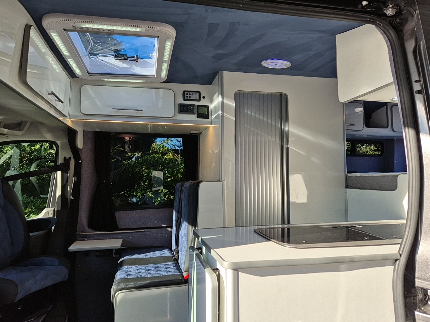 Inside a campervan showing seats, a window with curtains, a kitchen counter with a stove, and overhead storage compartments.