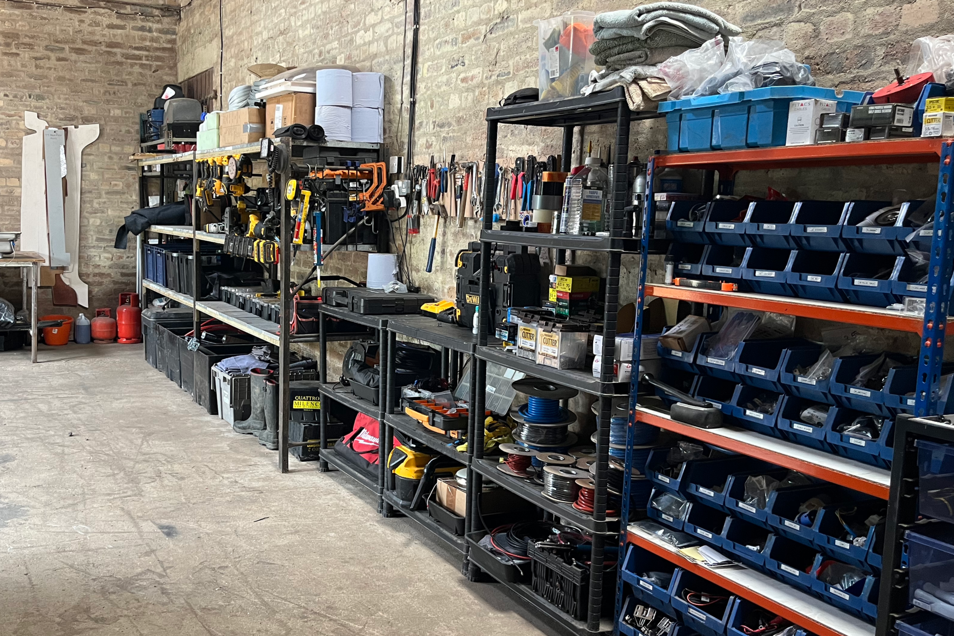 Workshop with organized shelves containing tools, equipment, and supplies. Visible shelving units hold blue bins, power tools, rewiring supplies, and various hardware items, with a brick wall background.