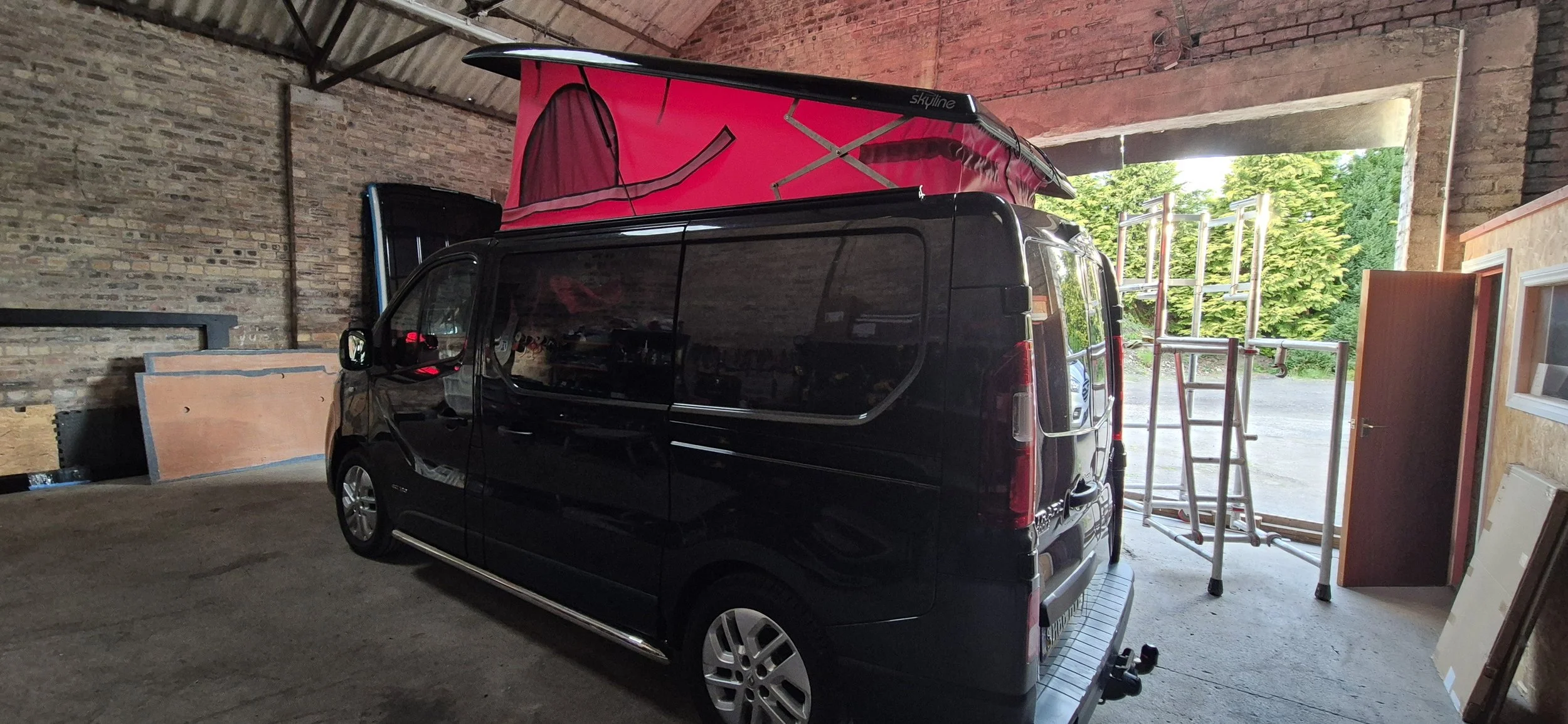 Black van parked inside a garage with a pink rooftop tent on top, surrounded by scaffolding and building materials, and open garage door revealing green trees outside.