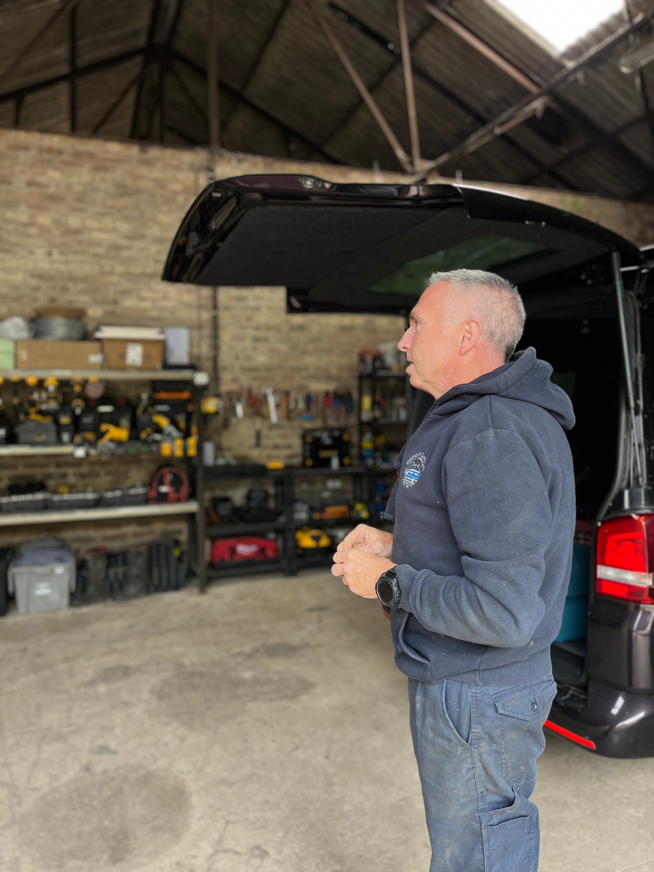 A man in a navy hoodie and jeans stands beside a black vehicle with an open trunk in a garage or workshop. Shelves with tools and storage boxes are visible in the background, and the structure has a brick wall and a high, sloped wooden roof.