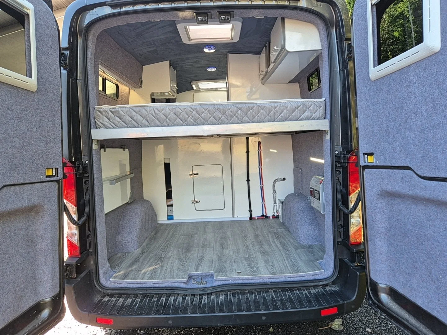 View of the inside of a converted van with a foldable bed, small storage cabinets, a wood floor, and various fixtures for camping or traveling.