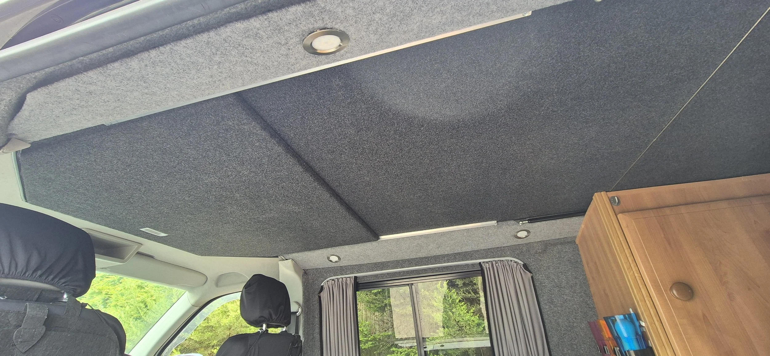 Interior of a vehicle with gray carpeted ceiling, gray curtains on window, and a wooden cabinet with books and items inside.