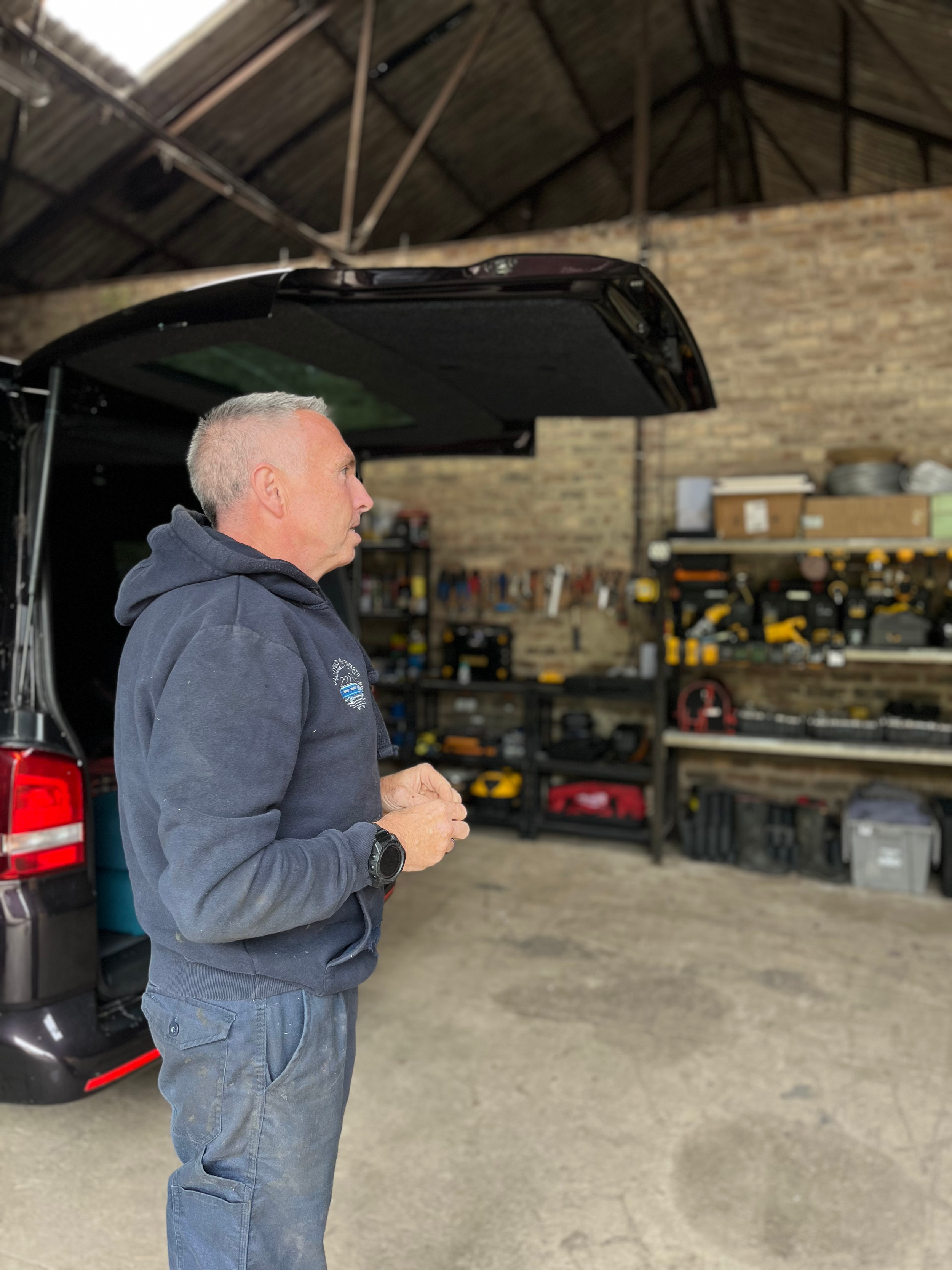A man stands in a workshop next to a black vehicle with an open trunk, inside shelves contain tools and equipment, with a brick wall in the background.