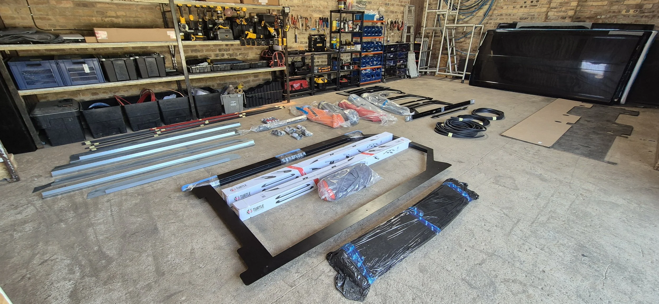 Garage or workshop with various tools, parts, and equipment organized on shelves, with a large black television or monitor, and numerous boxes and cables laid out on the floor.