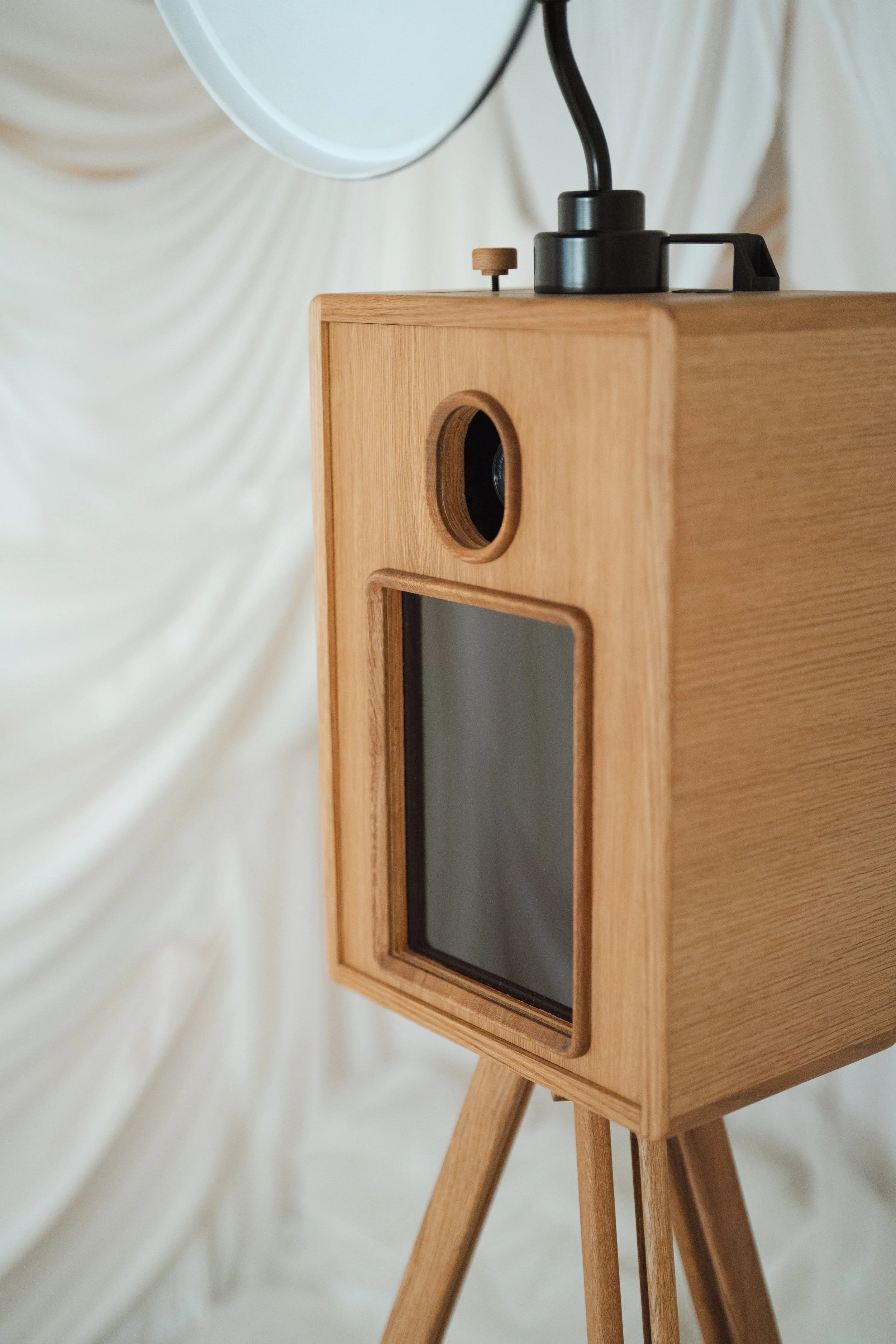 A wooden photo booth with a small lens and a screen, set on tripod legs, against a background of white draped fabric.