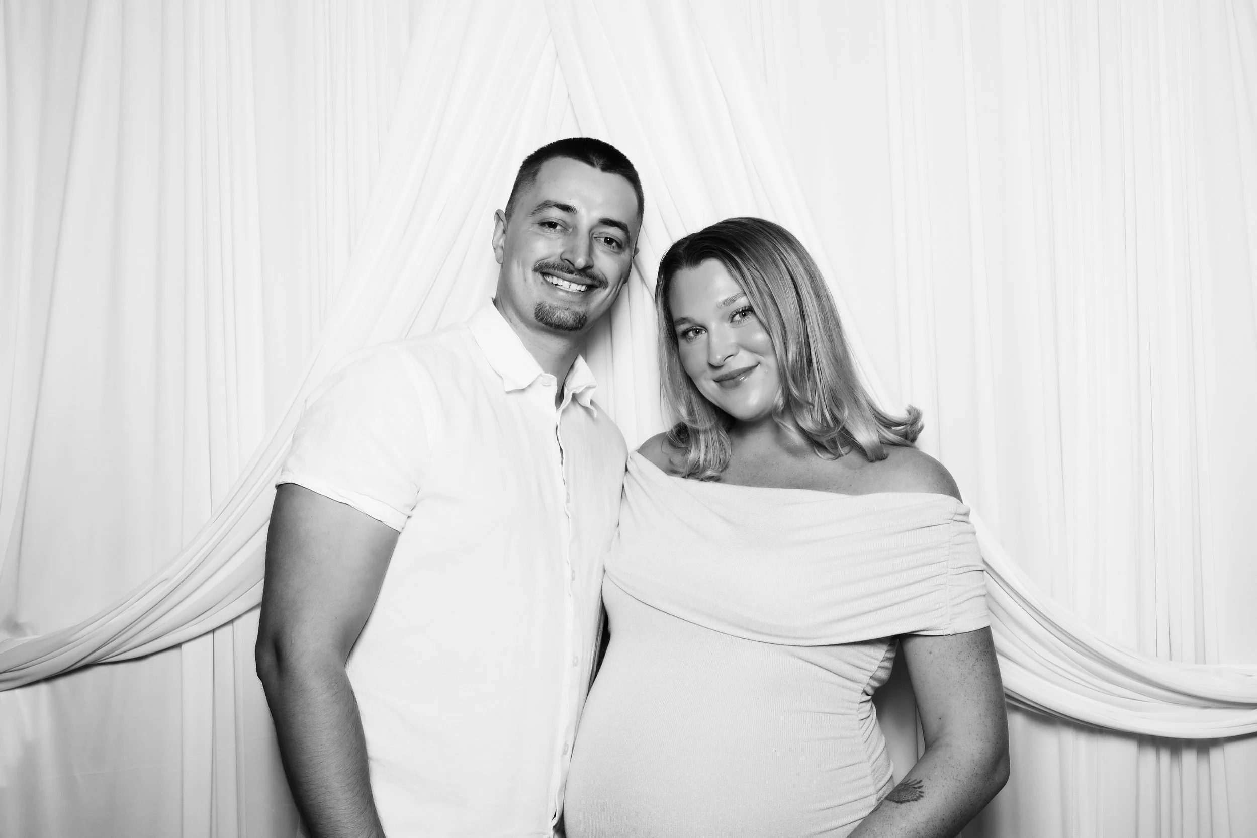A smiling man and woman posing together, standing in front of draped curtains. The man has short dark hair and a trimmed beard, wearing a short-sleeved shirt. The woman has shoulder-length blonde hair, wearing a fitted off-the-shoulder dress with a t