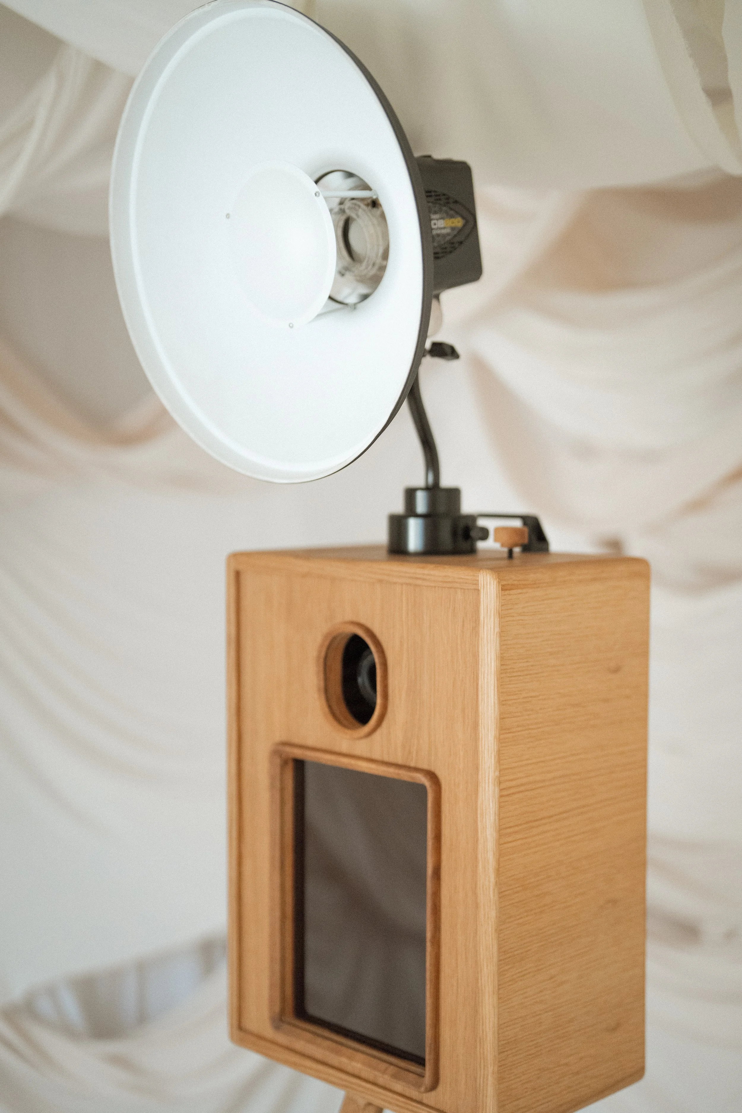 Photograph of a photo booth with a tripod, a large white circular reflector, and a wooden frame housing a camera and screen, set against a cream-colored draped fabric background.