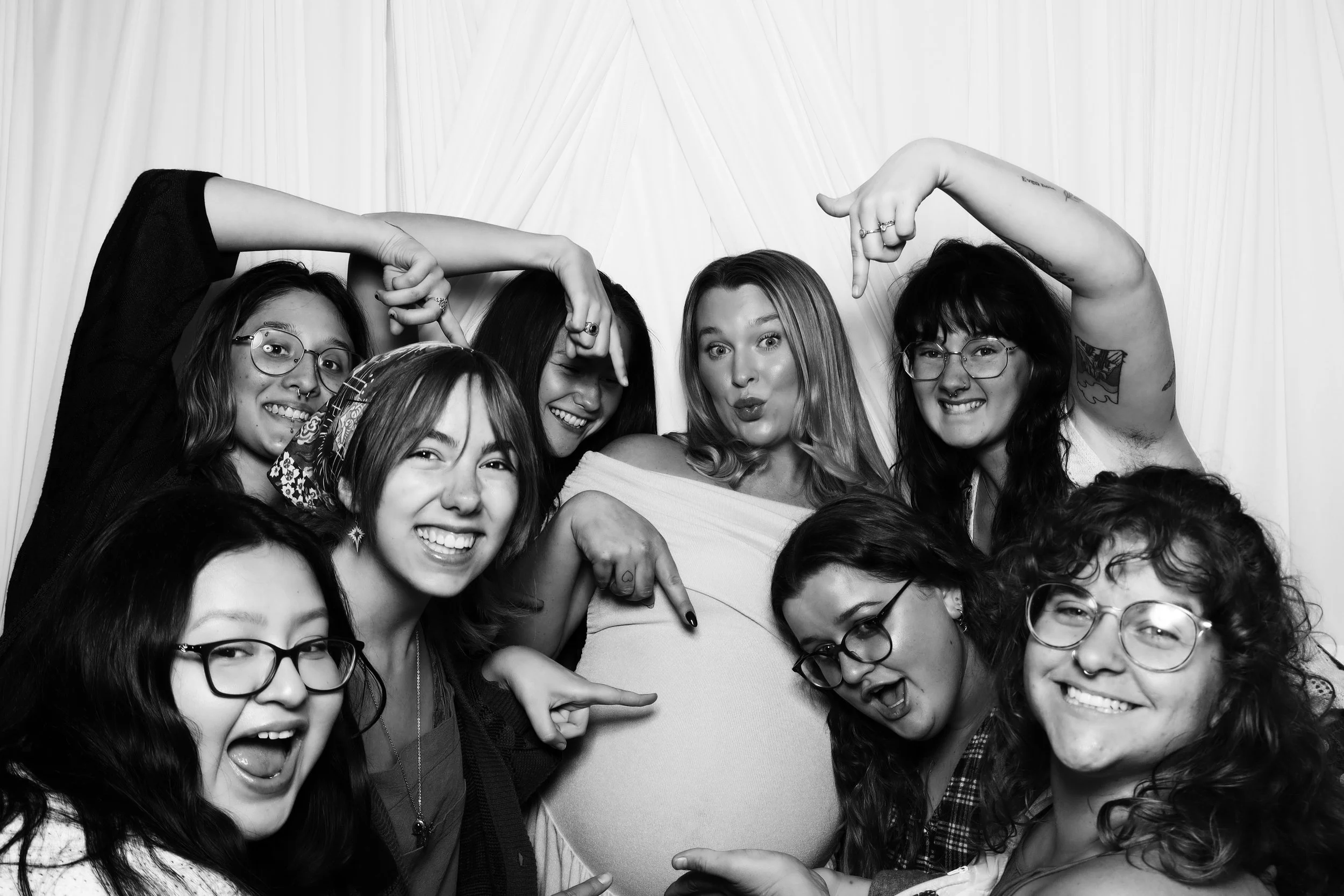 Group of smiling women posing together for a photo, some pointing at a large heart-shaped object in front of them.