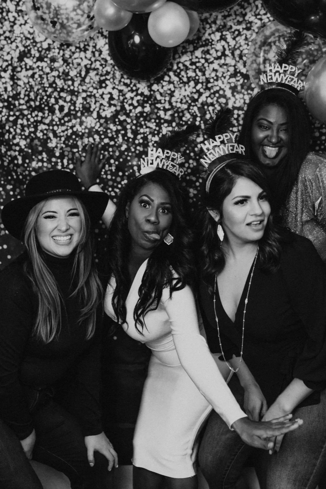 Four women celebrating New Year's Eve with party hats and balloons, smiling and posing for a photo in front of a dotted backdrop.