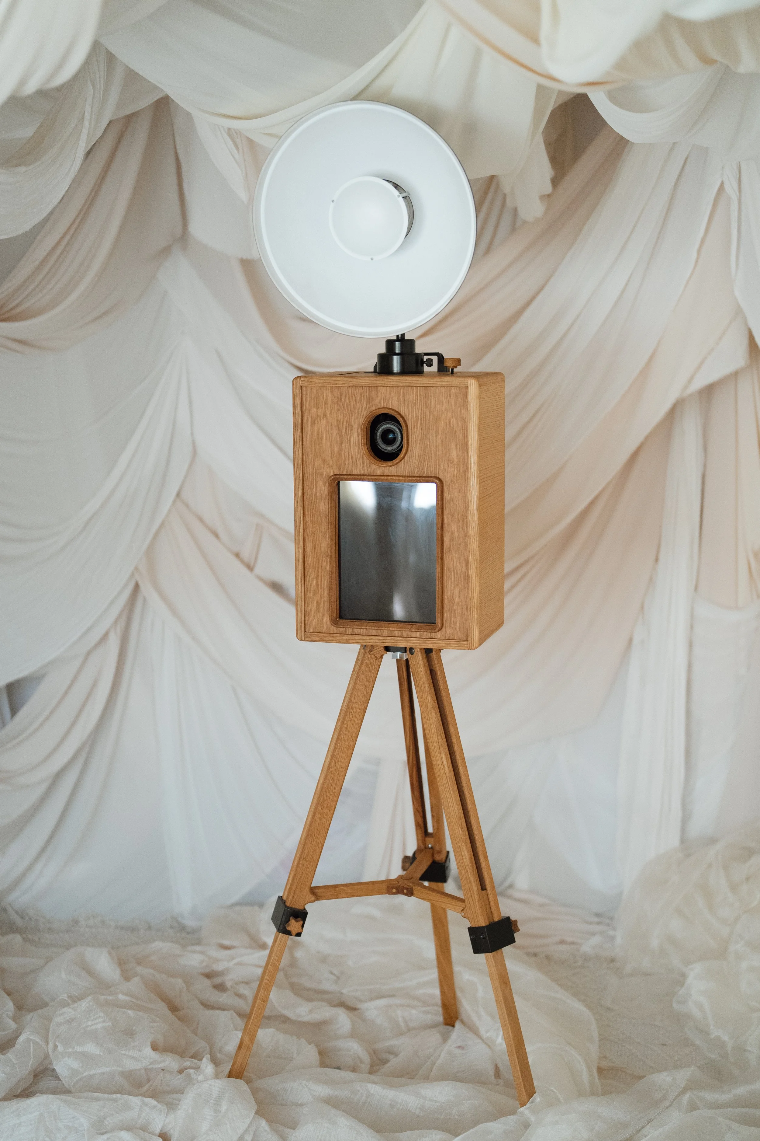 A vintage wooden photo booth camera on a tripod, with a white dome-shaped flash, set against cream-colored draped fabric background.