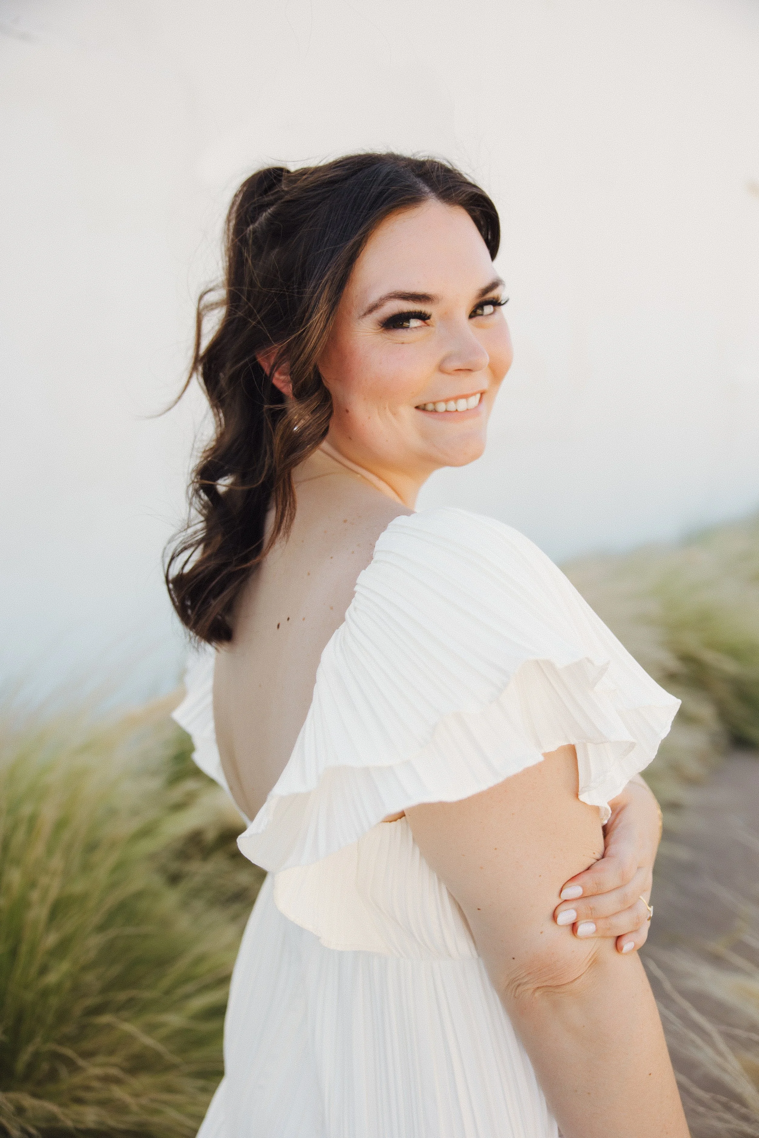 A woman with dark brown, wavy hair and light skin standing outdoors at a wedding, smiling over her shoulder, wearing a white dress with ruffled sleeves.