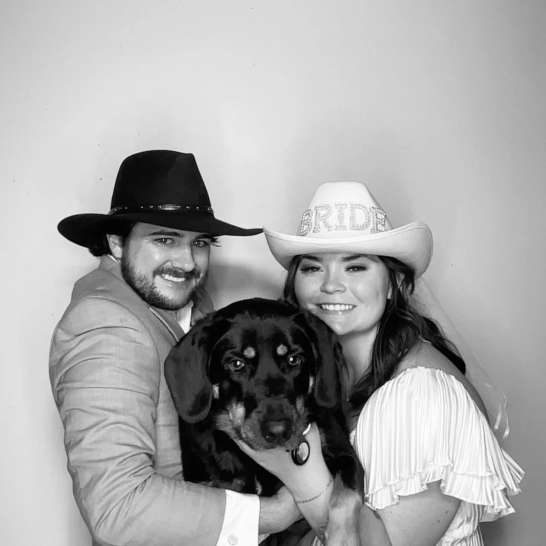 A black and white photo of a smiling couple holding a dog. The man is wearing a dark cowboy hat and a light-colored suit, while the woman is wearing a white cowboy hat with the word 'BRIDE' on it and a ruffled dress. The dog is a medium-sized, black and tan breed, looking at the camera.