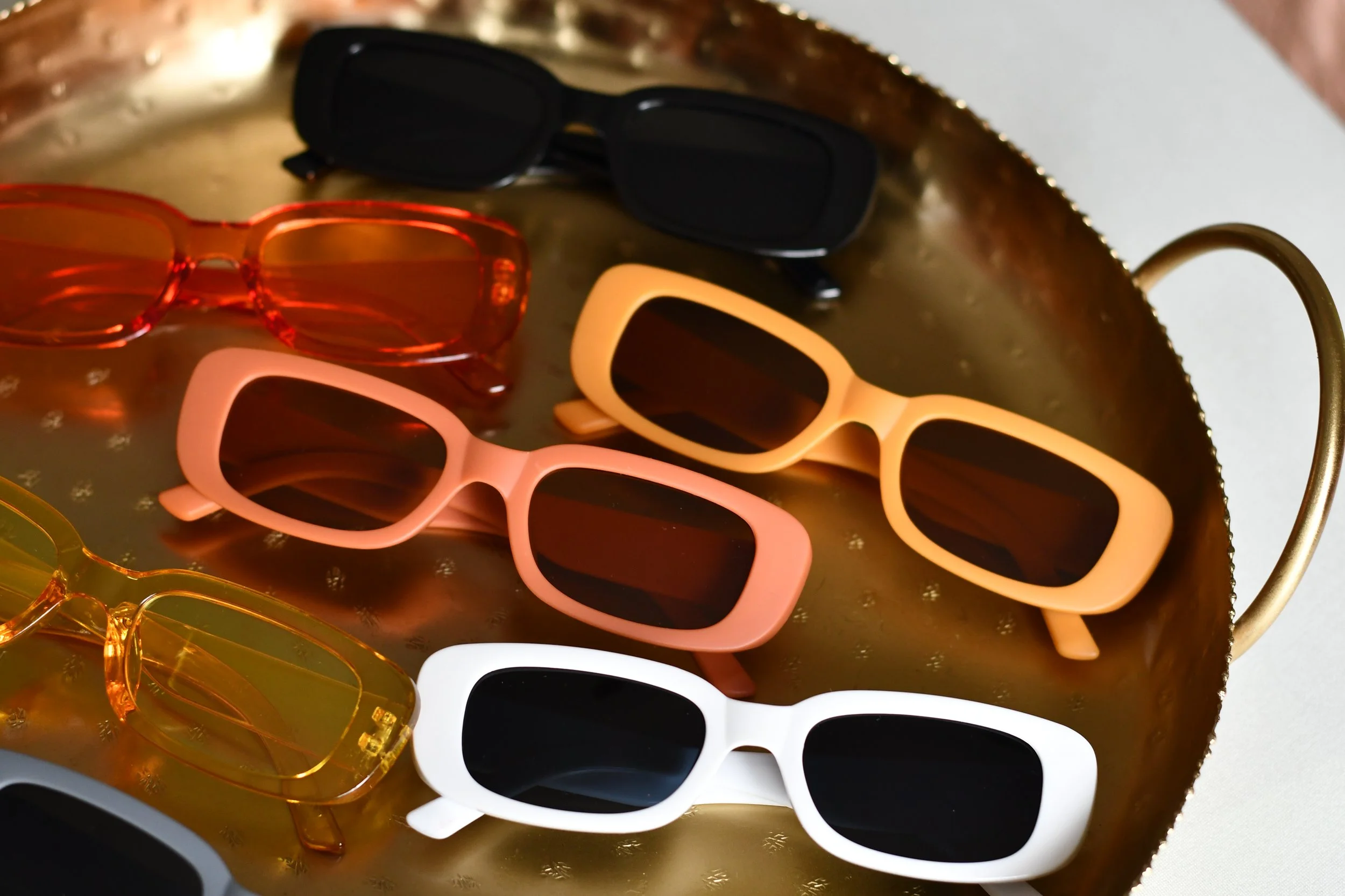 Six pairs of sunglasses with different frame colors—black, red, peach, yellow, white, and transparent—placed on a brass tray.