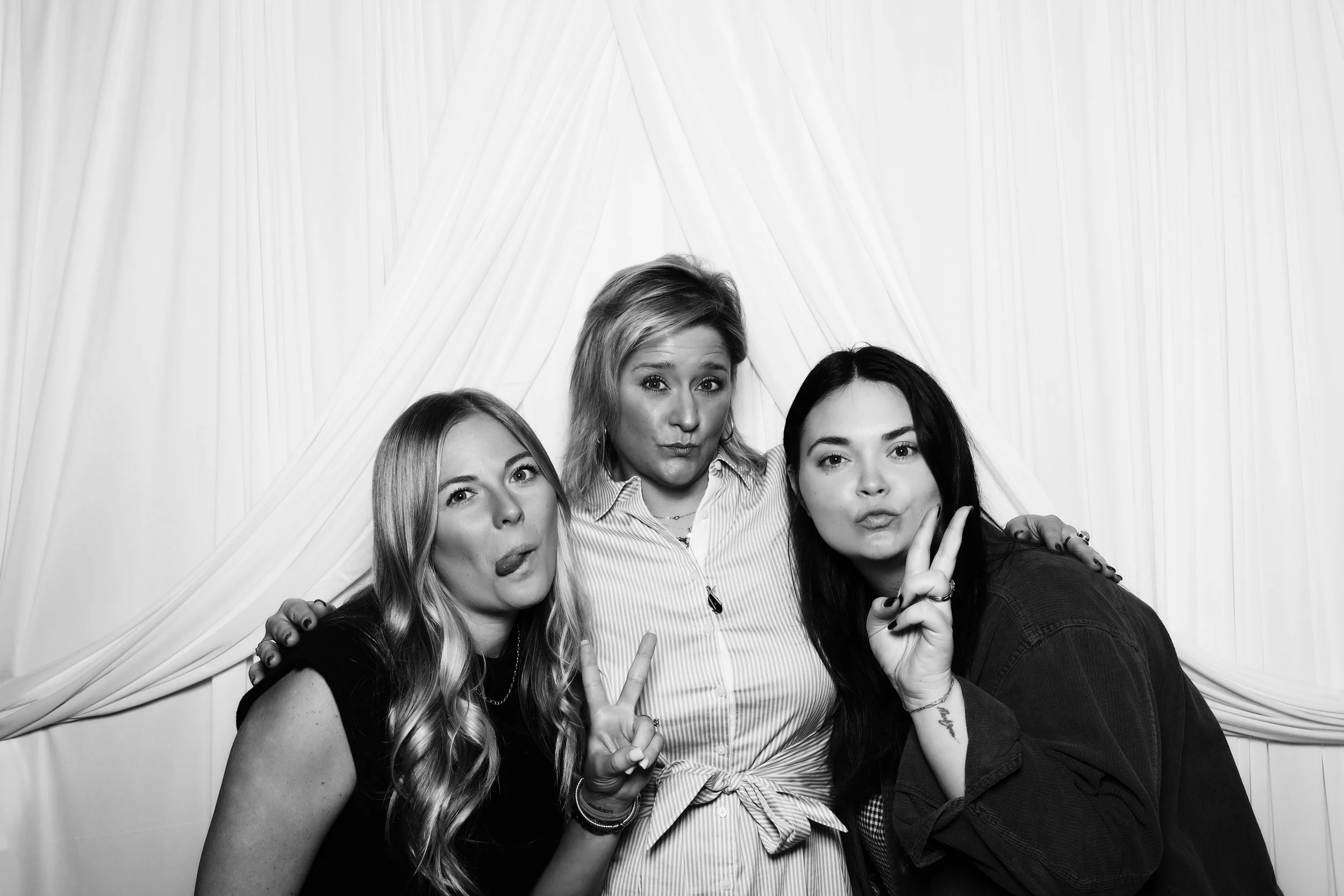 Three women posing playfully for a photo in front of a draped backdrop, with two making peace signs and sticking their tongues out.