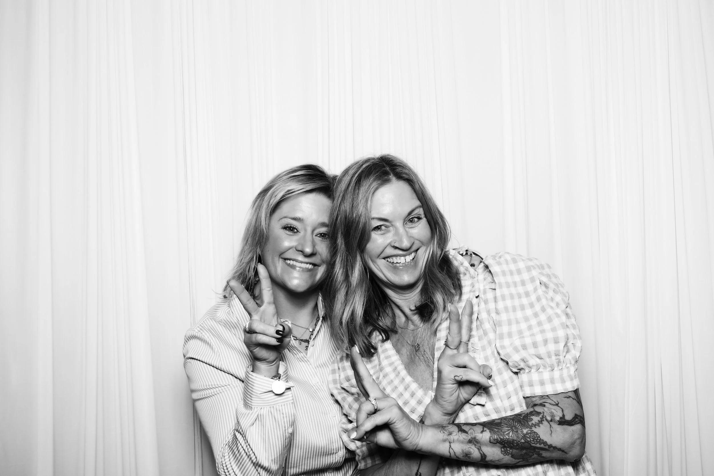 Two smiling women posing together, one making a peace sign with fingers, in front of a white curtain background.