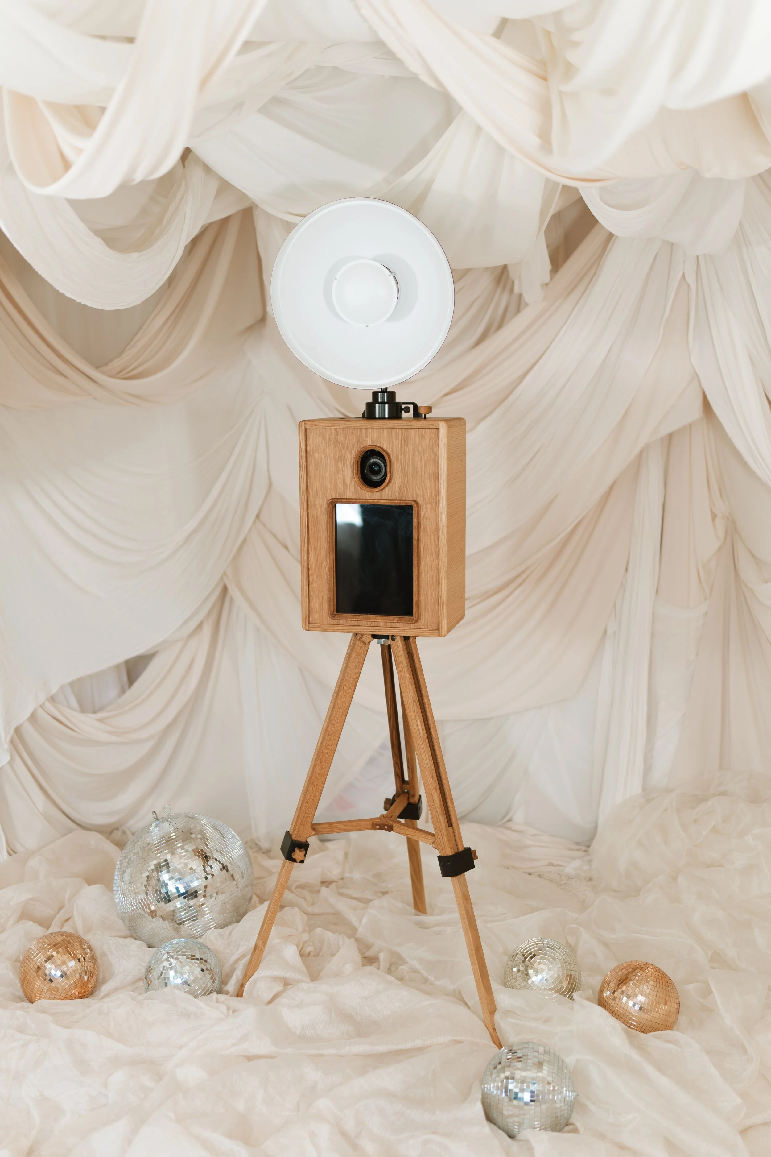 A photo booth with a wooden tripod, a camera, and a large white reflector, set against cream-colored drapery, with disco balls on the floor.